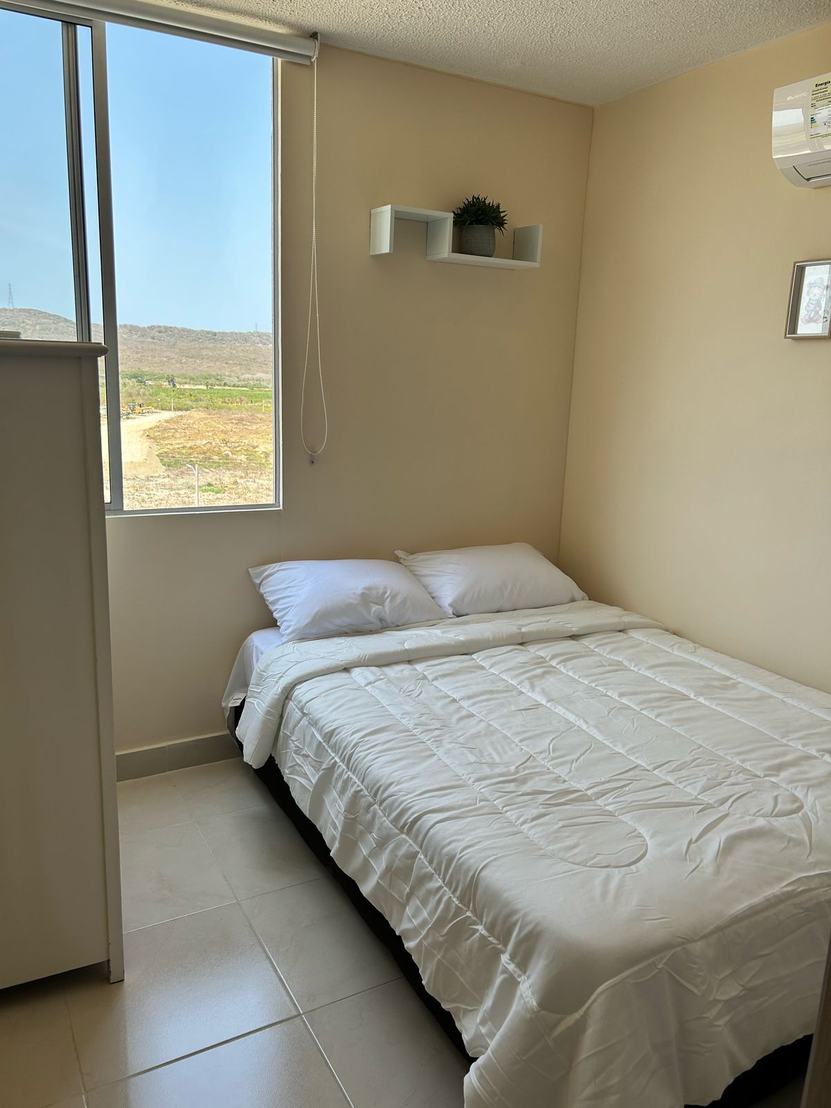 Een slaapkamer met een bed, een raam, een klein plankje met een plant en een luchtverkoeler aan de muur.