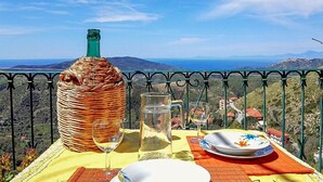 A table with a bottle, glasses, and plates on a balcony overlooking a scenic landscape.