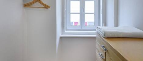Ein kleiner, sauberer Raum mit einem hölzernen Schminktisch, einem Fenster mit Blick auf Gebäude und einem hölzernen Kleiderhaken an der Wand.