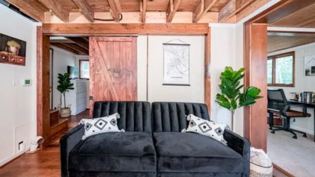 A cozy living room with a black sofa, wooden ceiling beams, a wooden door, and a framed map on the wall.