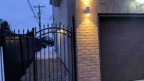 A gated driveway leading to a garage with a brick wall and a light fixture.
