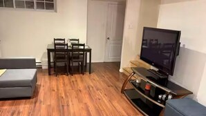 A living room with a grey sofa, a wooden dining table with chairs, a flat-screen TV on a stand, and a wooden floor.