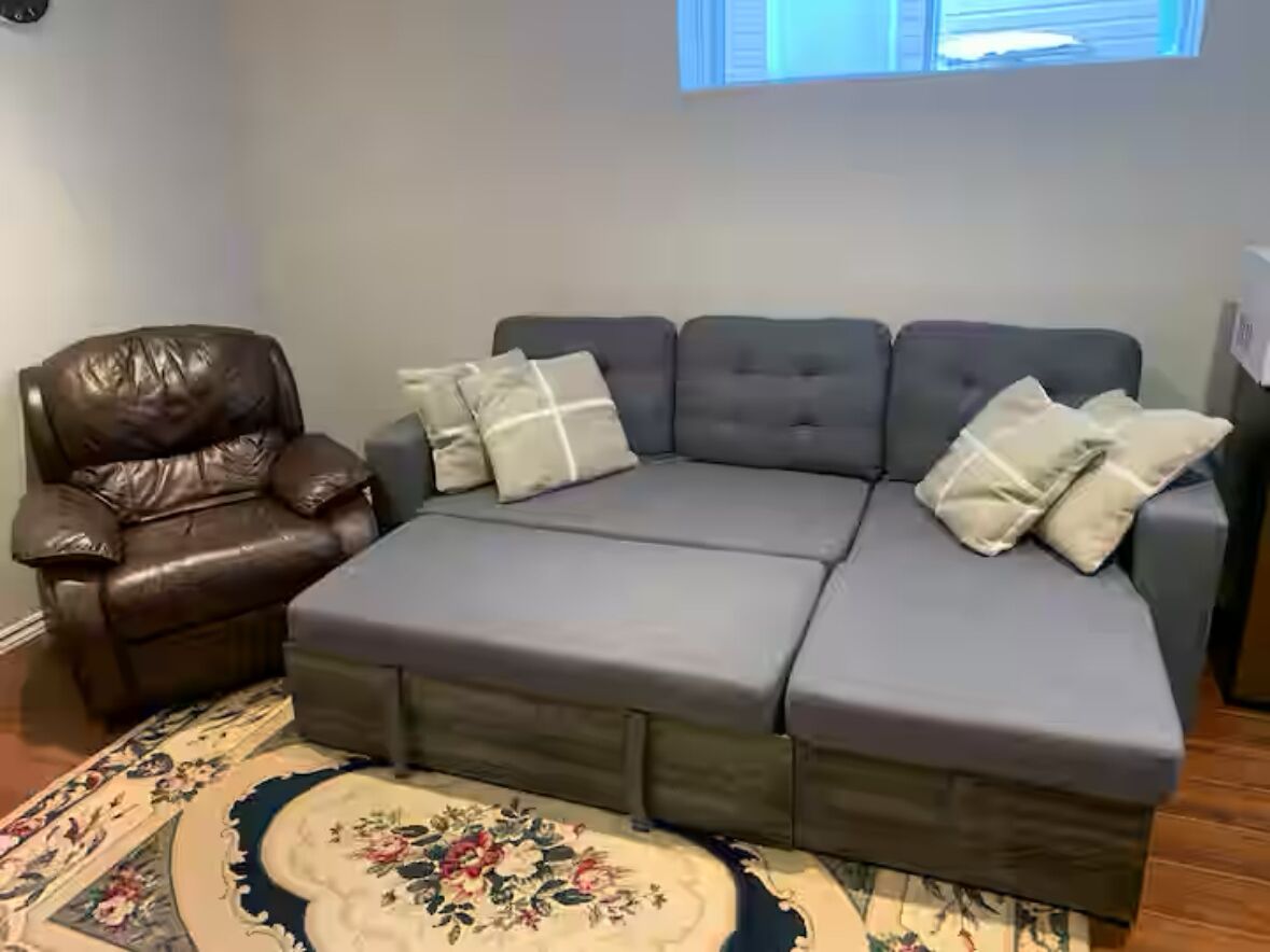 Un sofá modular con tela gris y un sillón de cuero marrón, una alfombra estampada y una ventana con persianas.