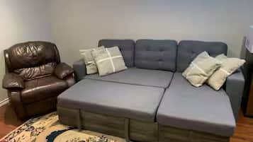 Un sofá modular con tela gris y un sillón de cuero marrón, una alfombra estampada y una ventana con persianas.