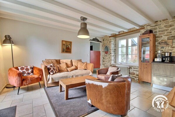 Una sala de estar acogedora con un sofá, sillones y una mesa de centro. Se aprecia una pared de piedra, una ventana y una zona de cocina con estufa.