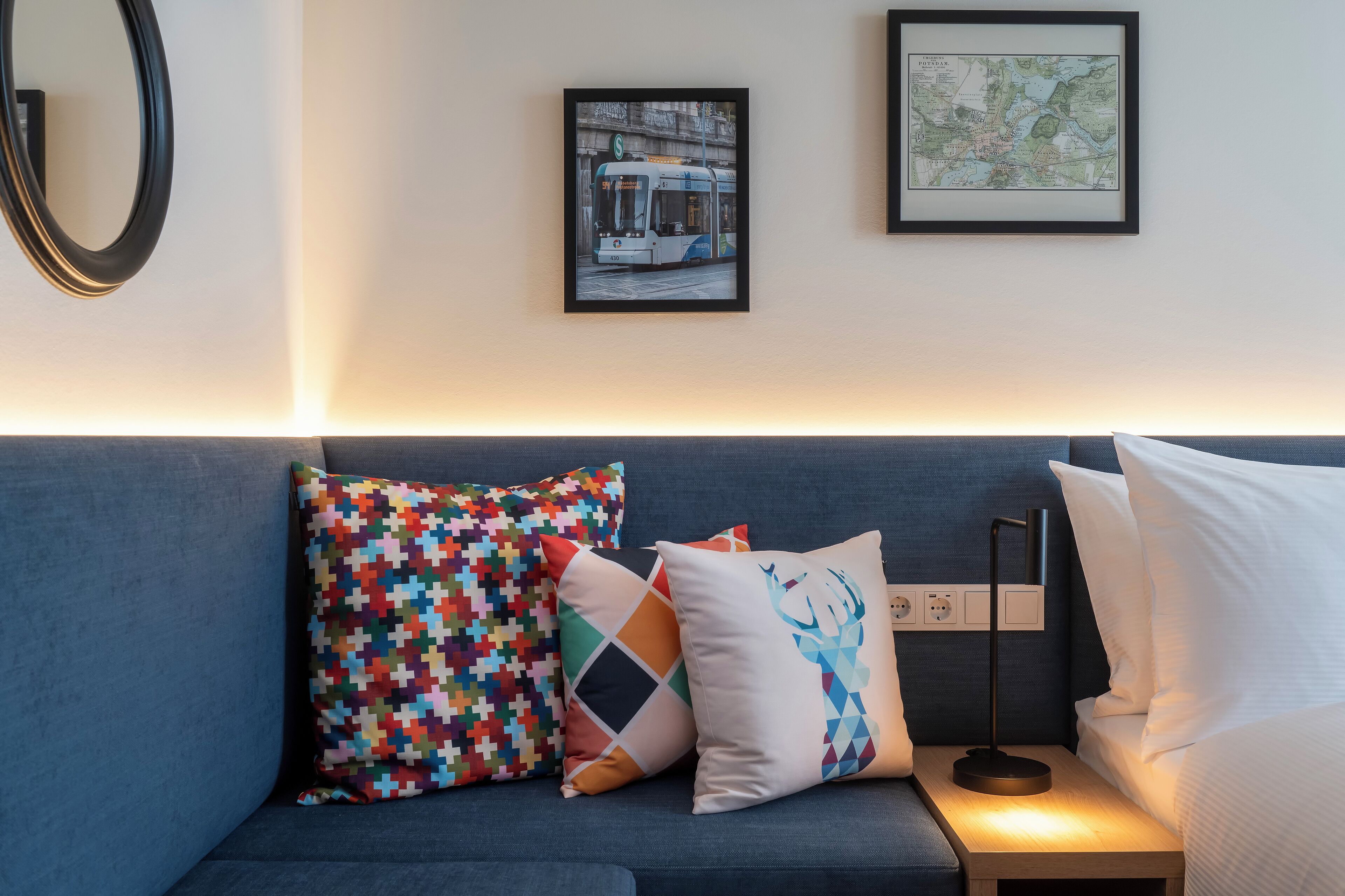 A modern hotel room with a blue sofa, colorful pillows, a framed cityscape picture, and a map on the wall.