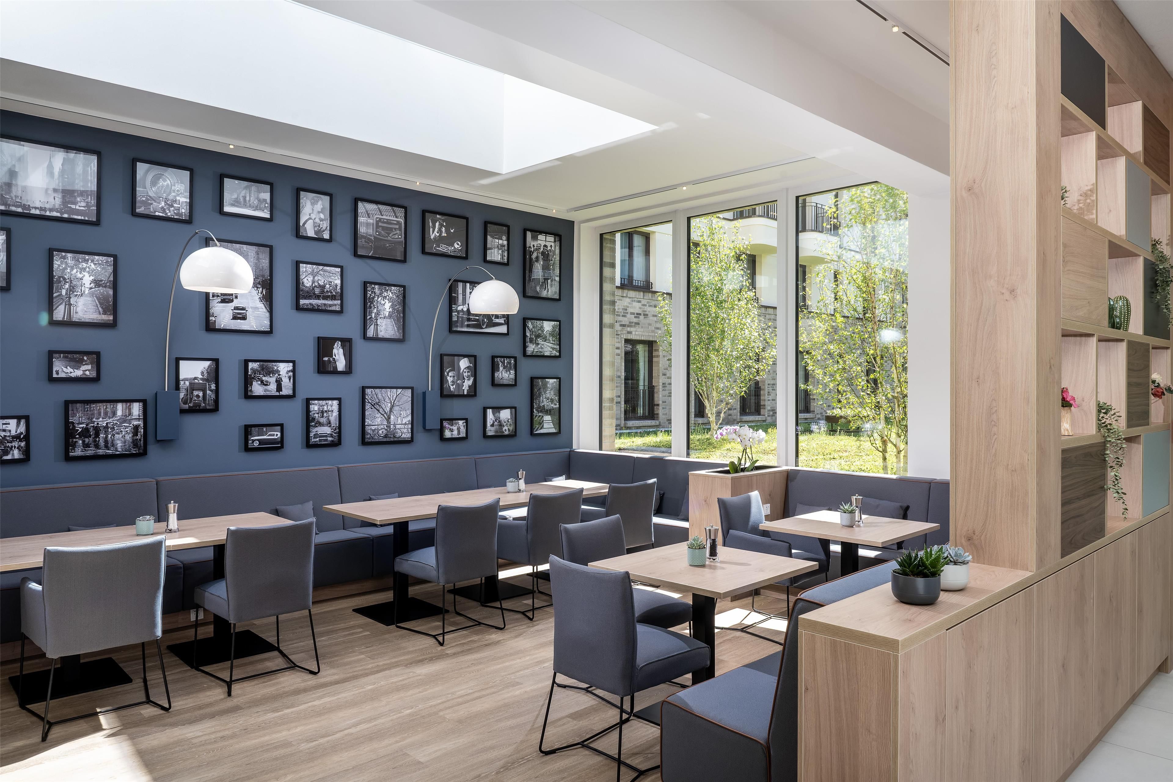 A modern restaurant with a blue wall adorned with black and white photographs, wooden tables and chairs, and a view of greenery through large windows.