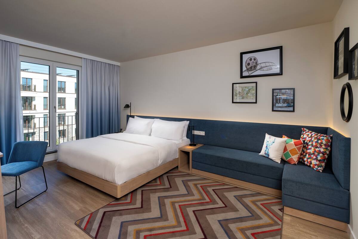 A modern hotel room with a large bed, a blue sofa, and a view of the city through the window.