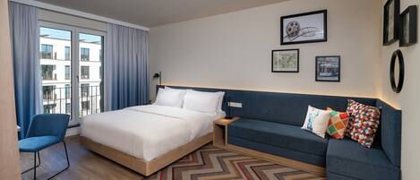 A modern hotel room with a large bed, a blue sofa, and a view of the city through the window.