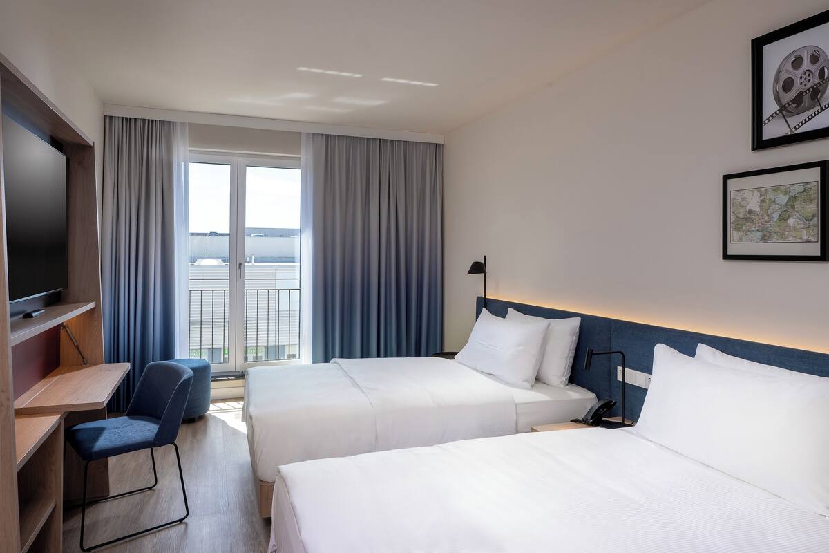 A hotel room with two beds, a desk, a chair, a TV, and a balcony with curtains.