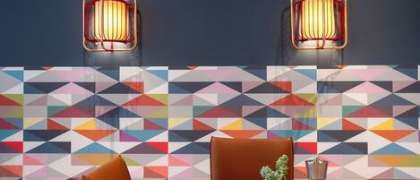 A dining area with a wooden table, two brown chairs, a potted plant, and a salt and pepper shaker. The wall features a colorful geometric pattern.