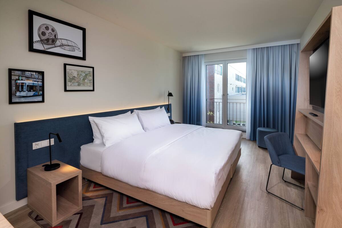 A modern hotel room with a large bed, a desk with a lamp, a TV, and framed pictures on the wall.
