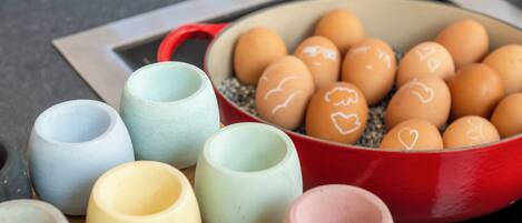 Una olla roja llena de huevos, algunos con dibujos de corazones, y varias tazas de cerámica pequeñas de distintos colores.