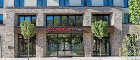 A modern hotel entrance with a brick facade, large glass doors, and a sign reading "Hampton by Hilton".