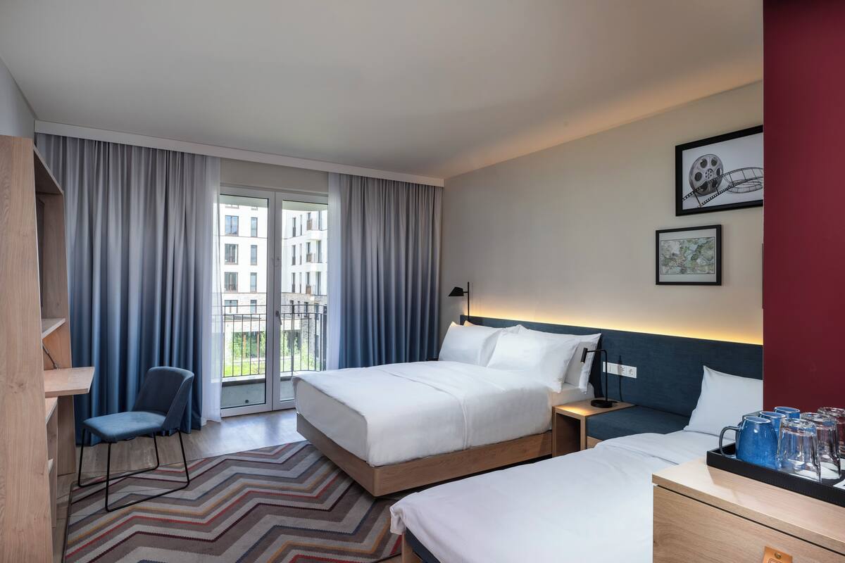A hotel room with a large bed, a smaller bed, a desk, a chair, and a view of the outside.