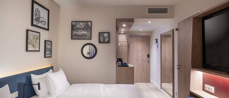 Habitación de hotel con cama, escritorio, televisor y espejo. Hay cuadros enmarcados en la pared y una puerta que da a otra habitación.