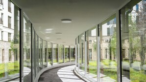 Un couloir aux murs de verre et au plafond incurvé, qui mène à un espace extérieur agrémenté de verdure, avec un bâtiment visible au loin.