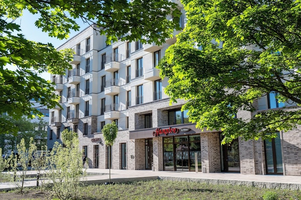 A modern building with a sign reading "Hampton by Hilton." It features multiple balconies and large windows.