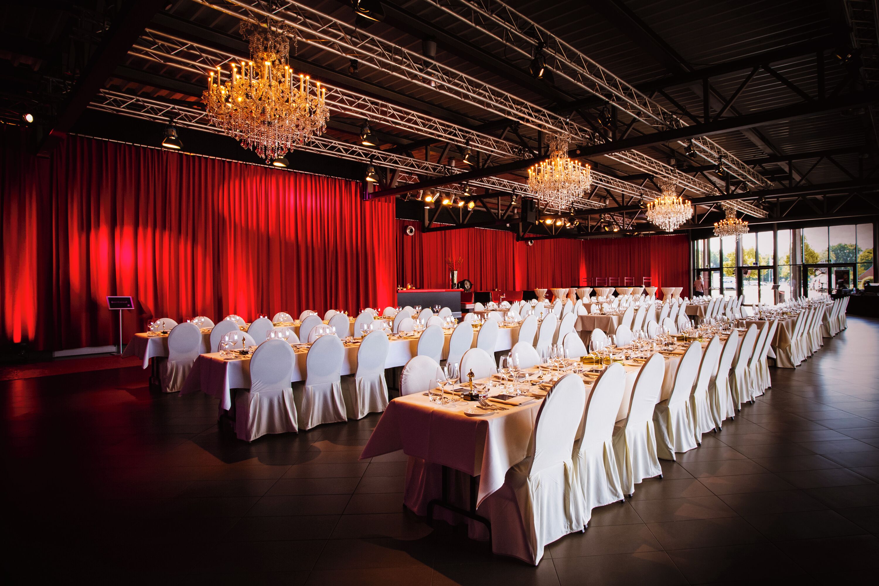 Un salón de banquetes con filas de mesas dispuestas para un evento formal, cortinas rojas, candelabros y un escenario.