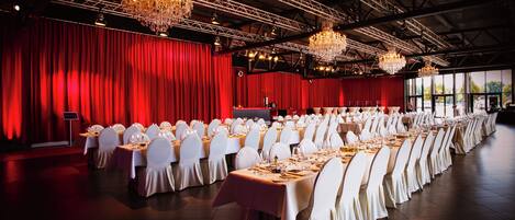 A banquet hall with rows of tables set for a formal event, red curtains, chandeliers, and a stage.