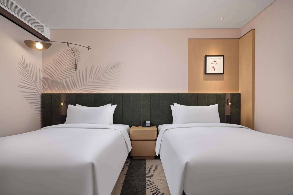 A hotel room with two beds, a nightstand, a framed picture on the wall, and a ceiling light fixture.
