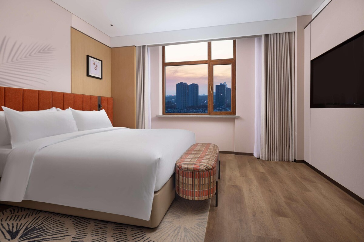 A hotel room with a large bed, a view of the cityscape through a window, a flat-screen TV, and a plaid ottoman.