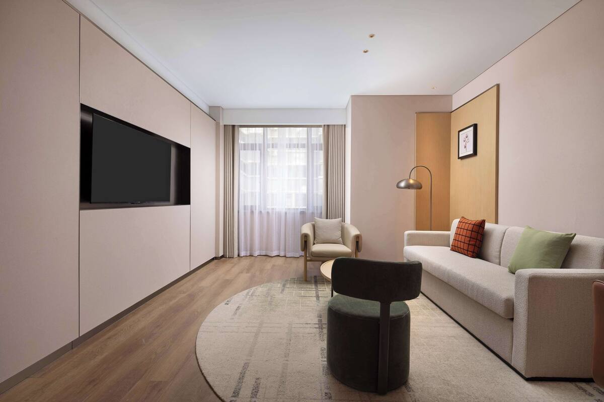 A modern living room with a flat-screen TV mounted on the wall, a round rug, a sofa with cushions, a chair, and a floor lamp.