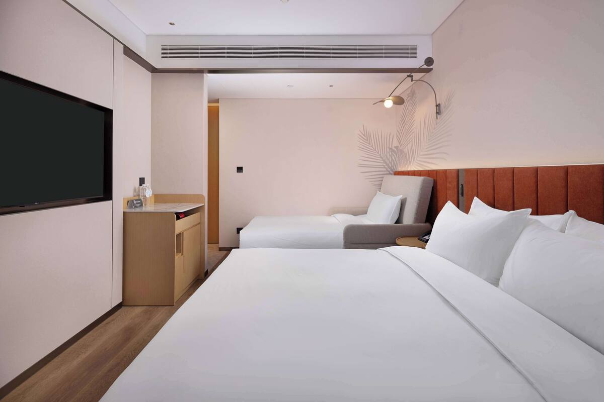 A hotel room with a large bed, a smaller bed, a desk with a chair, a TV, and a wall-mounted lamp.