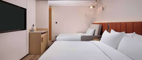 A hotel room with a large bed, a smaller bed, a desk with a chair, a TV, and a wall-mounted lamp.