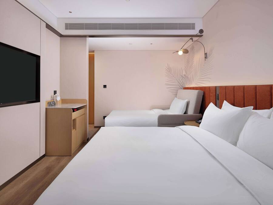 Habitación de hotel con una cama grande, una cama más pequeña, un escritorio con silla, un televisor y una lámpara de pared.