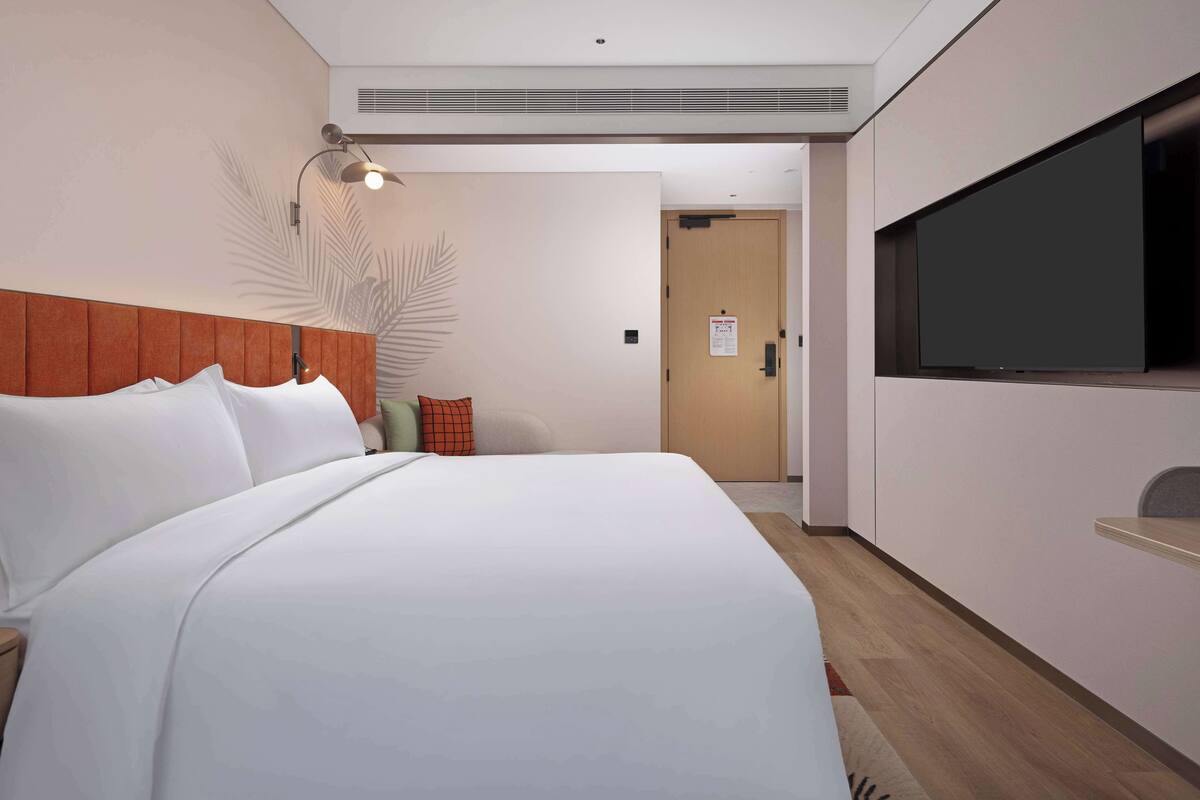 A hotel room with a large bed, a flat-screen TV mounted on the wall, a wooden headboard, and a wall-mounted lamp.