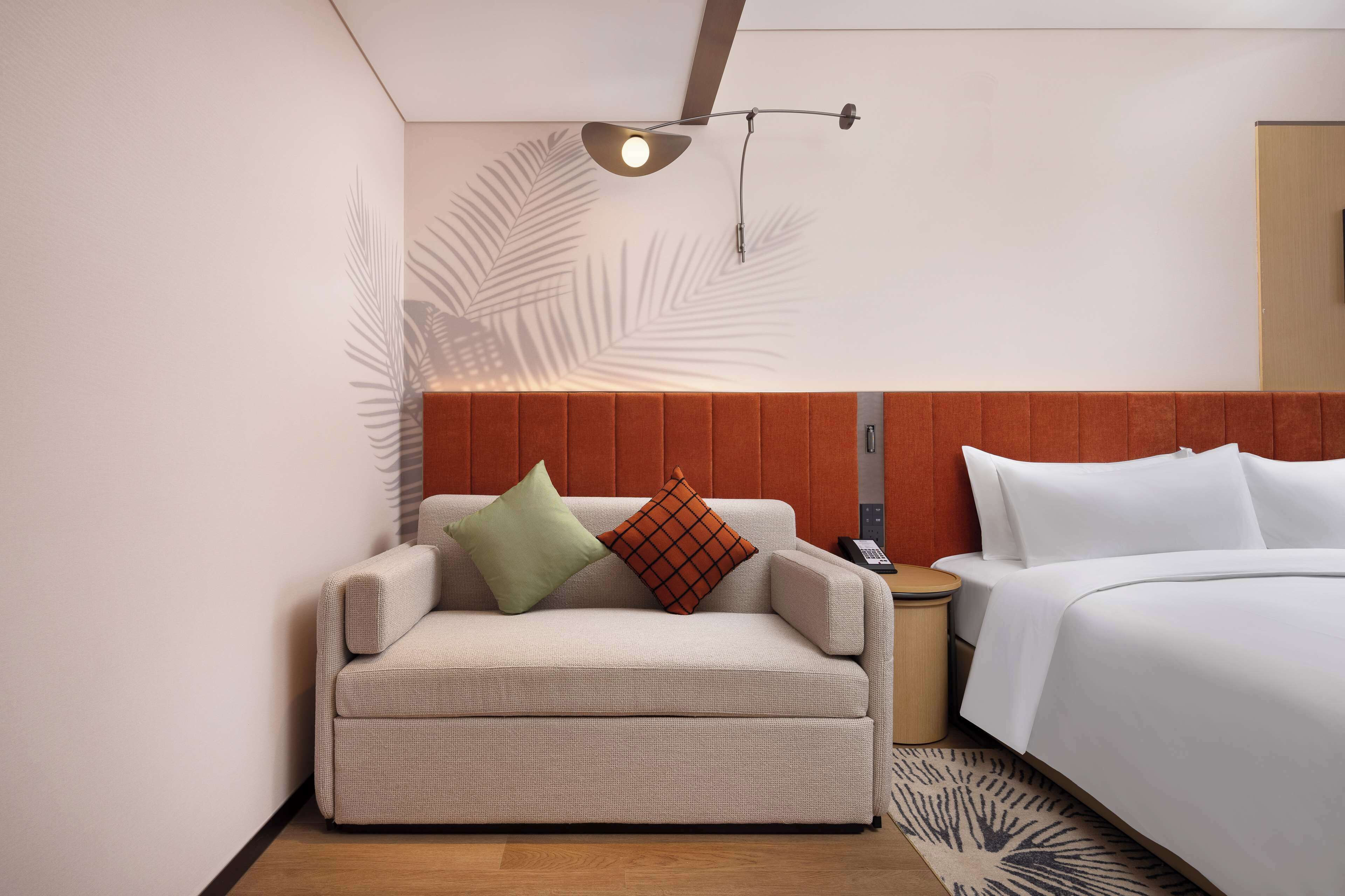 A hotel room with a sofa, a bed, a nightstand, and a wall-mounted lamp.