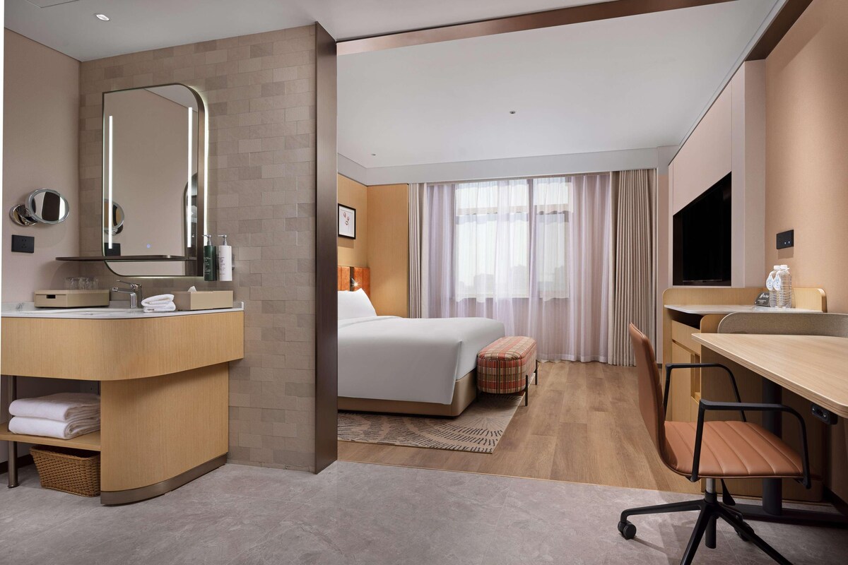 Une chambre d’hôtel moderne dotée d’un grand lit, d’un coin salon avec un bureau et une chaise, d’une coiffeuse avec un miroir et d’une télévision.