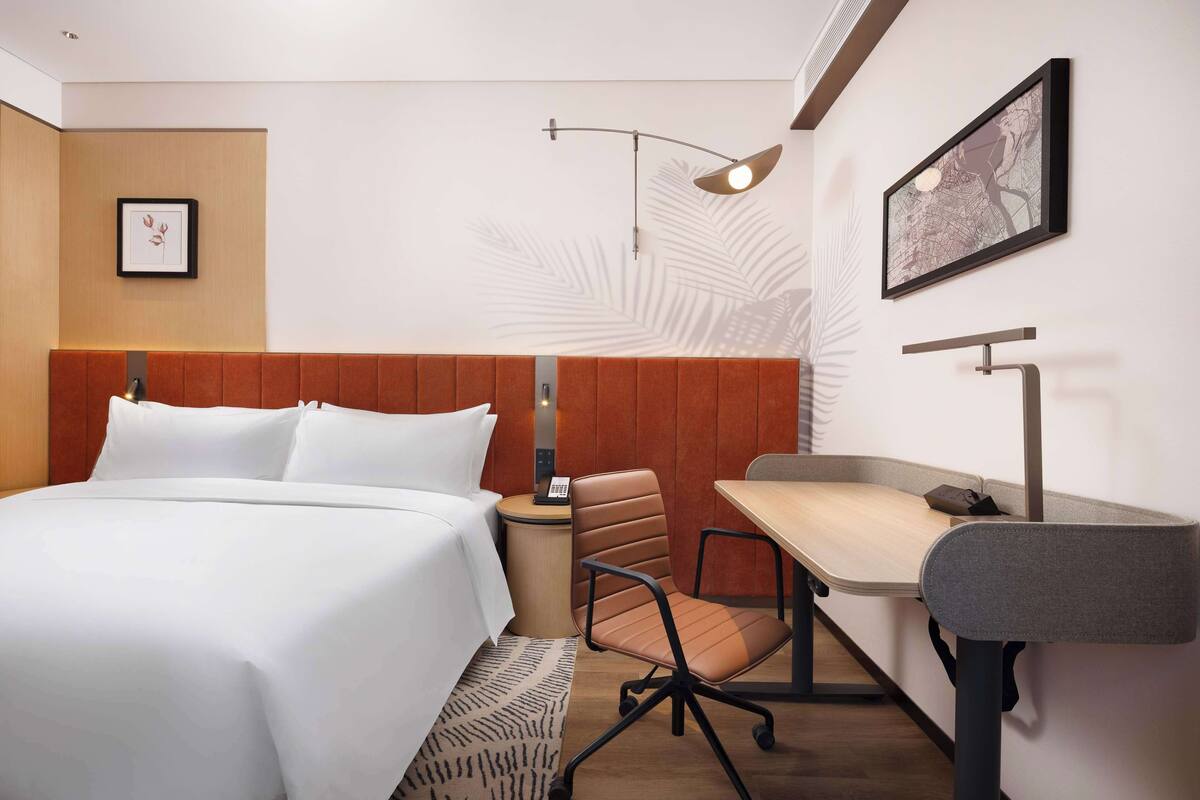 A hotel room with a bed, a desk, a chair, a lamp, and a framed map on the wall.