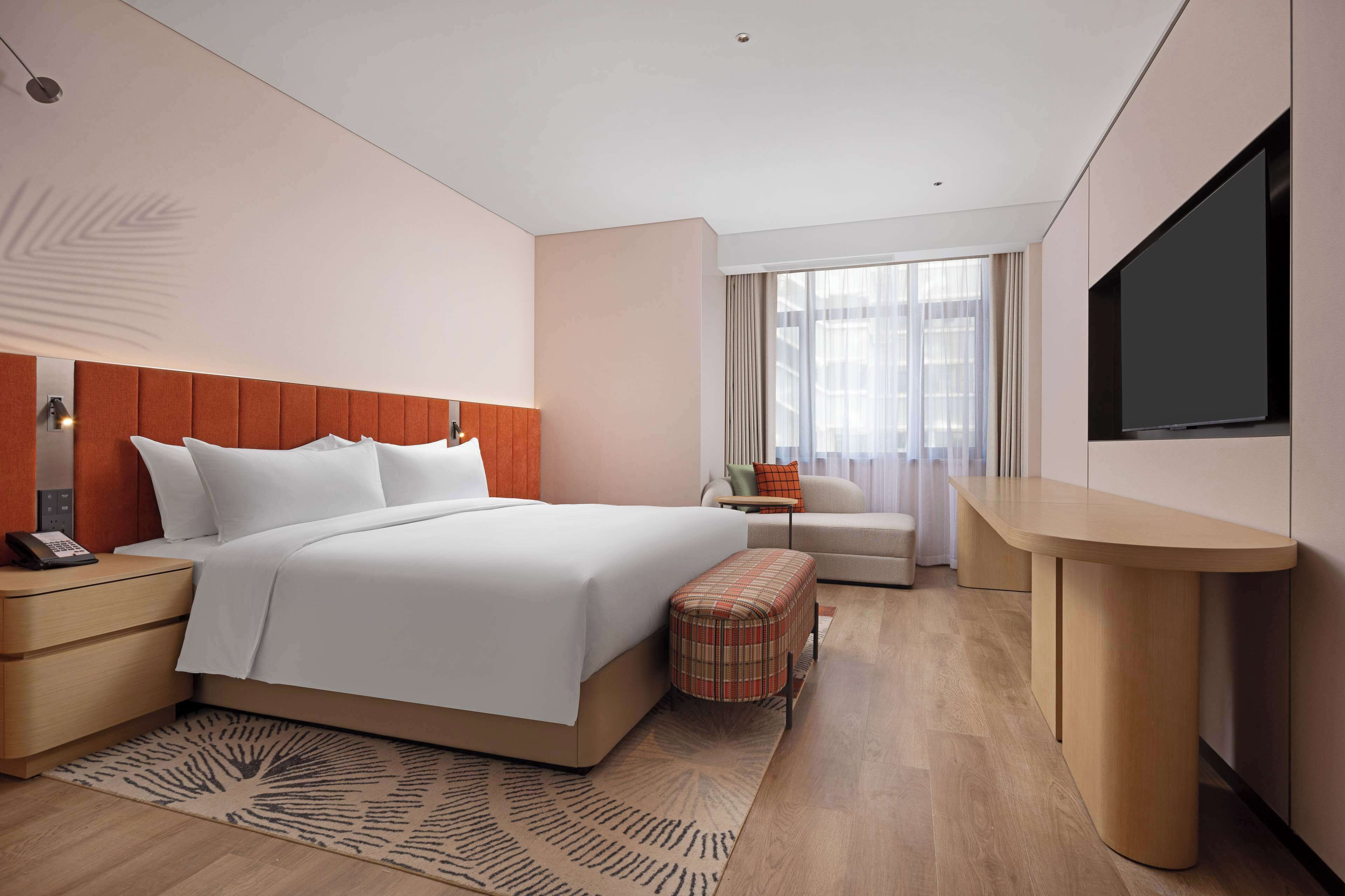 Ein modernes Hotelzimmer mit einem großen Bett, einem Nachttisch mit Telefon, einem kleinen Hocker, einer Couch, einem an der Wand befestigten Fernseher und einem großen Fenster mit Vorhängen.