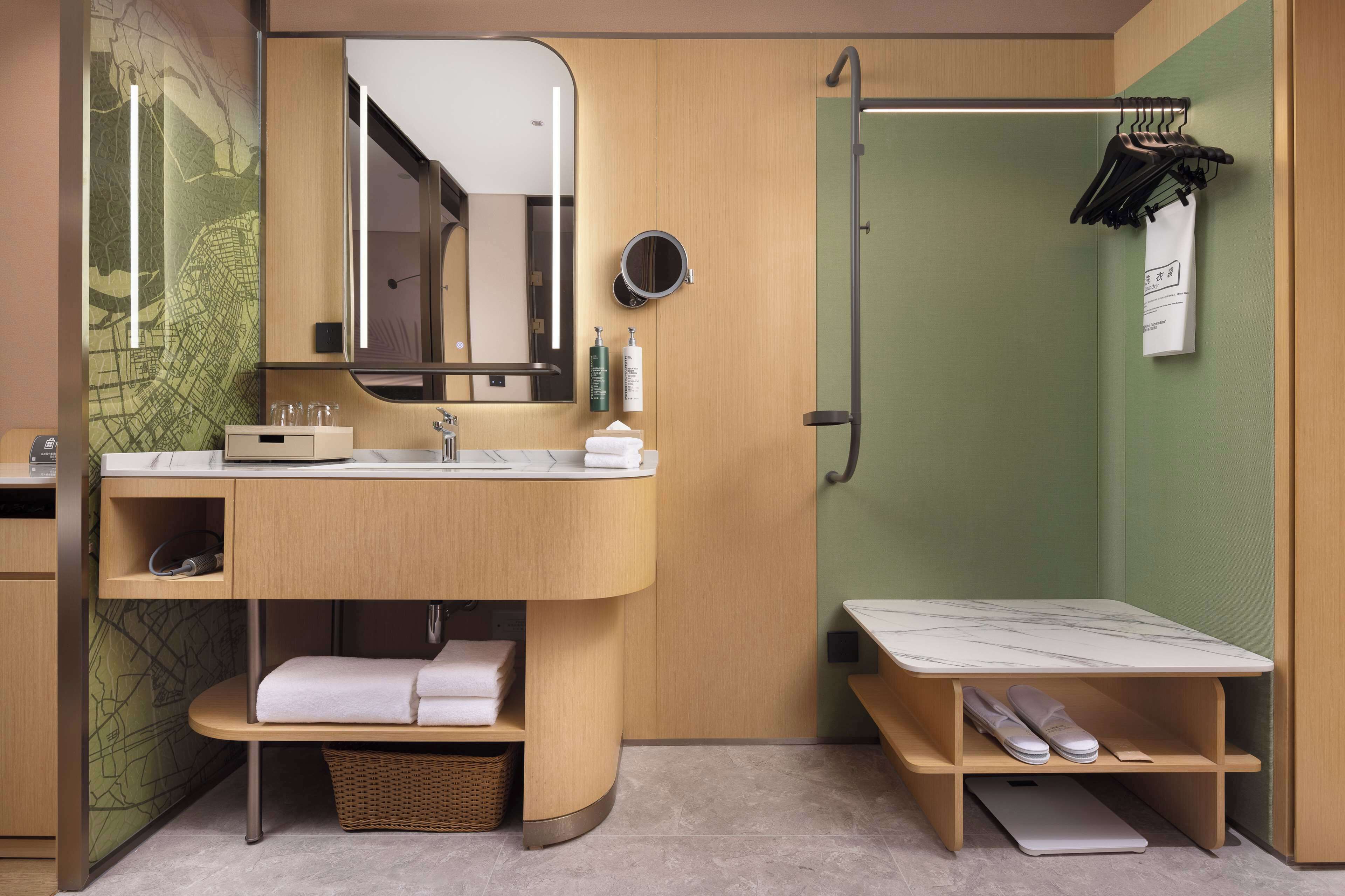 A modern bathroom with a curved vanity, a mirror, a shower, and a small seating area with a laptop and slippers.