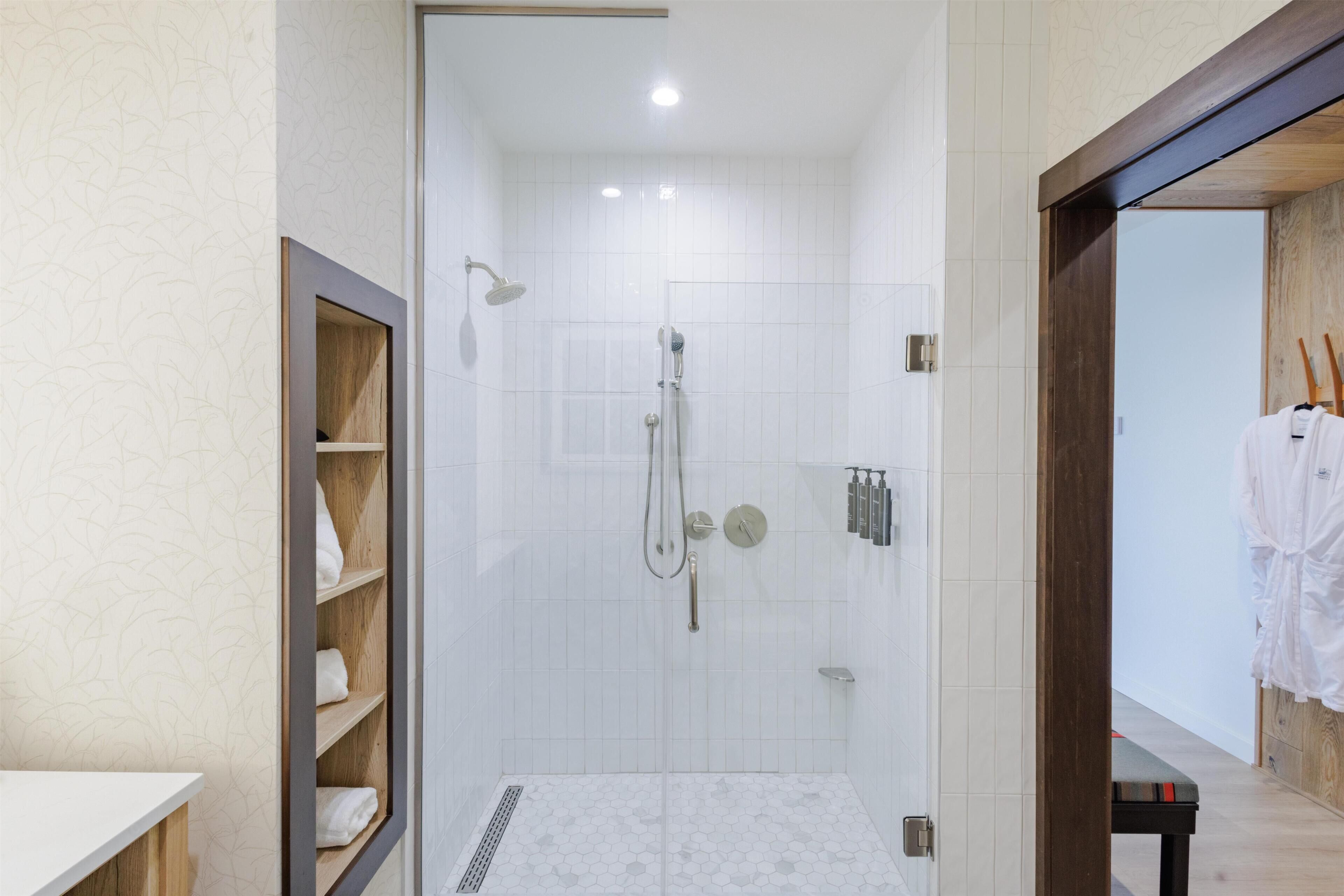 A modern bathroom with a shower, a wooden shelf with towels, and a white tiled floor.