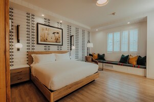 A bedroom with a wooden bed, a wall-mounted picture, a bedside table, a bench with cushions, and a window with blinds.