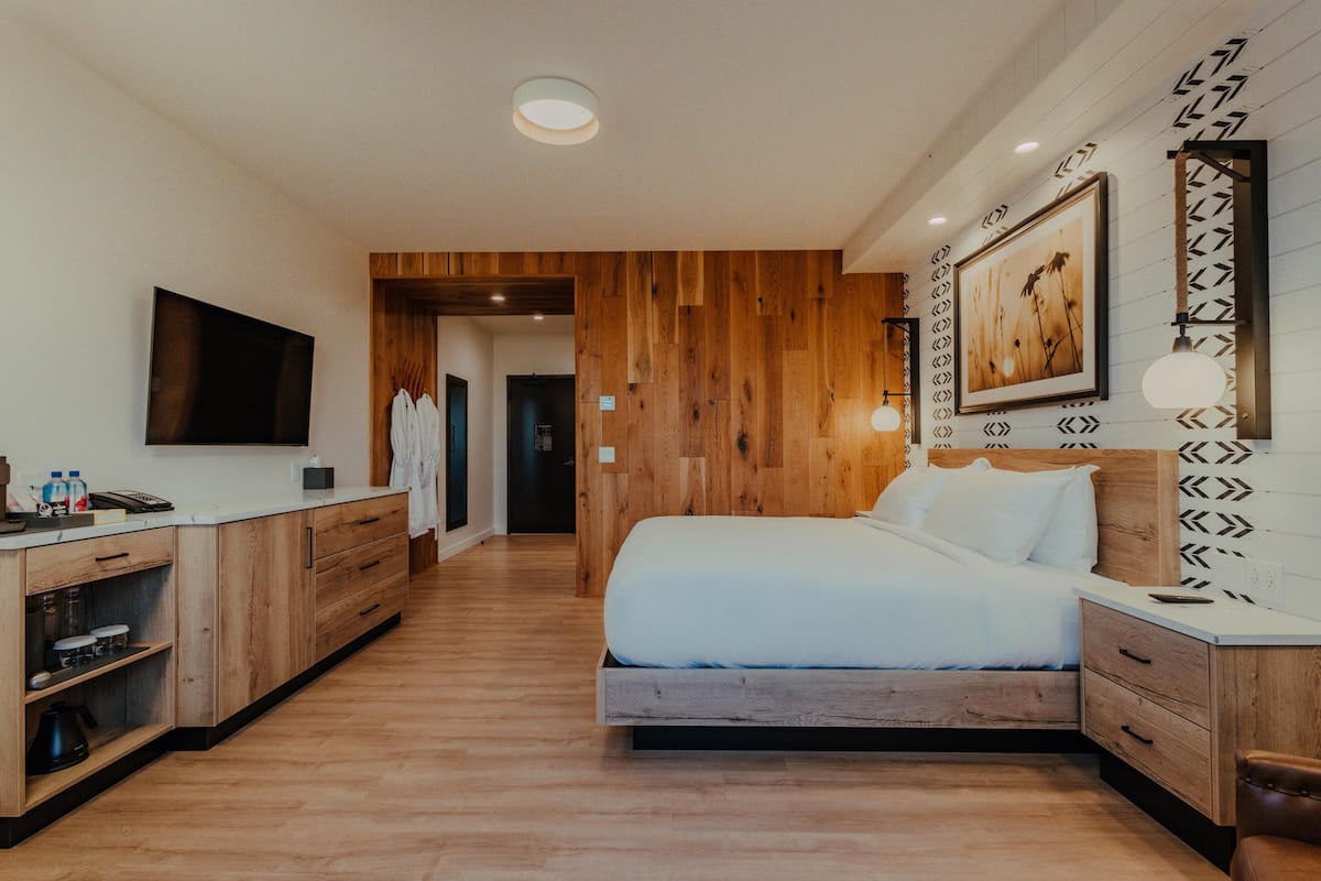 Un dormitorio moderno con suelo de madera, una cama con ropa de cama blanca, un escritorio con televisor de pantalla plana y un cuadro colgado en la pared.