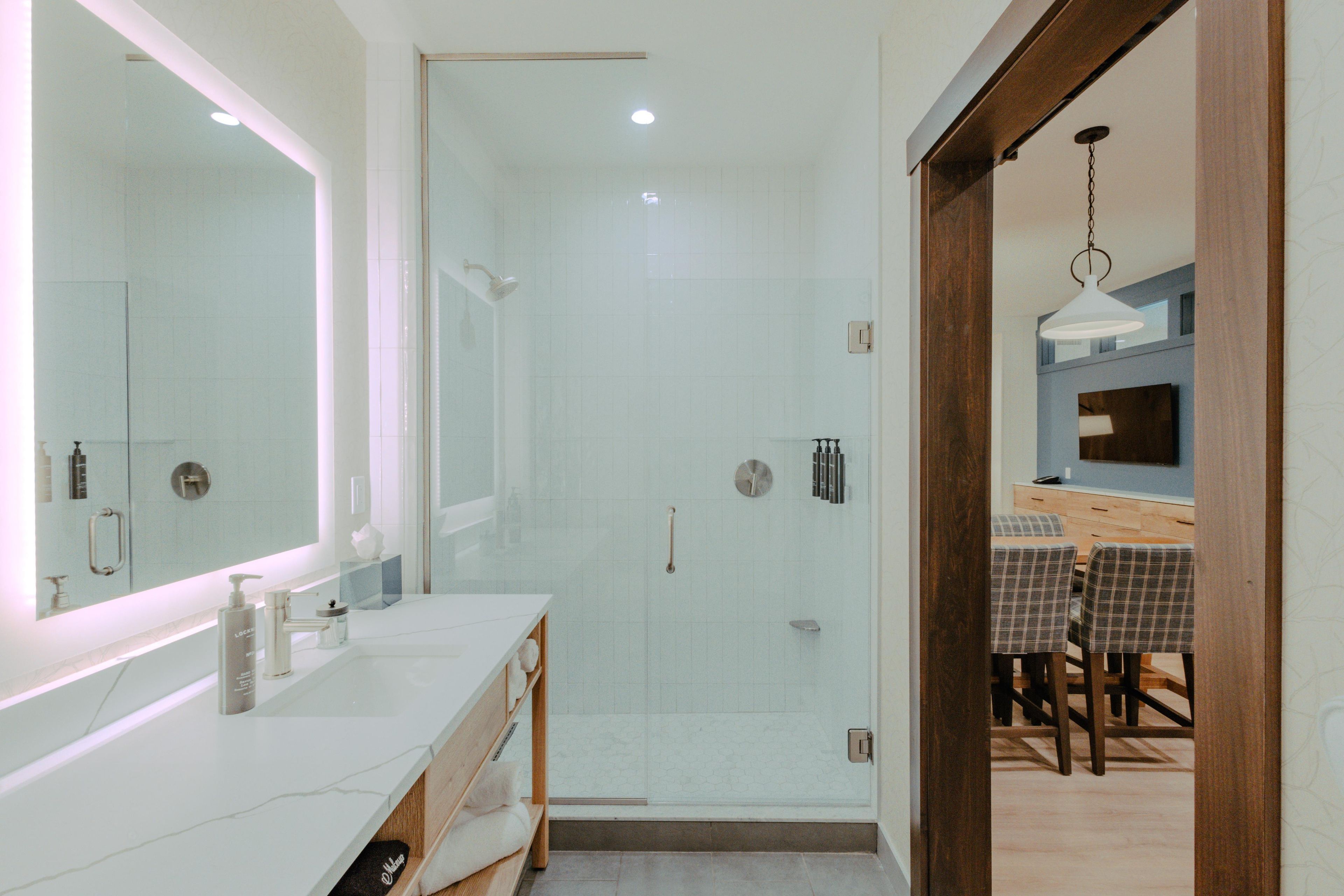 A modern bathroom with a glass shower enclosure, a vanity with a mirror, and a dining area visible through an open doorway.
