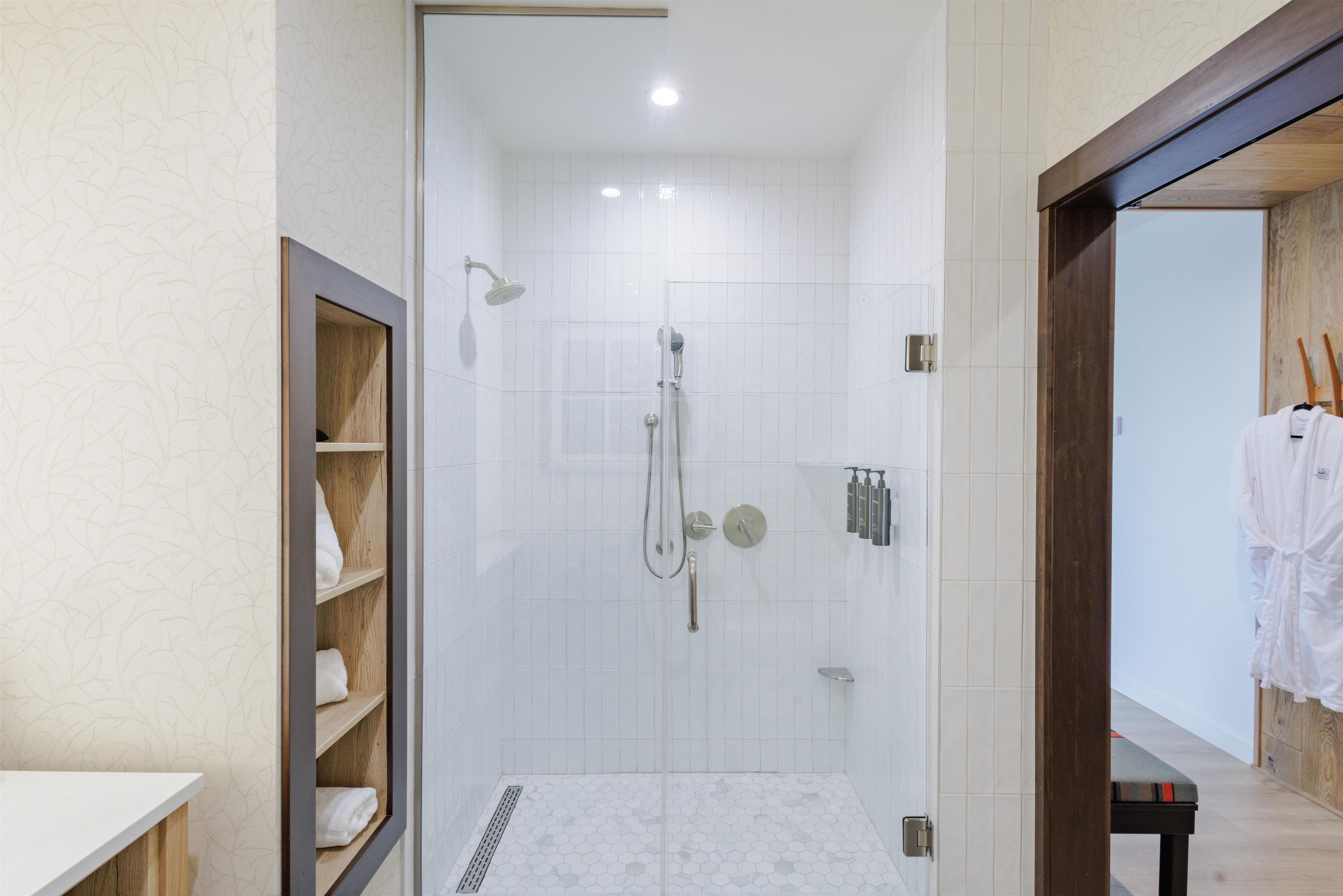 A modern bathroom with a shower, a wooden shelf with towels, and a white tiled floor.
