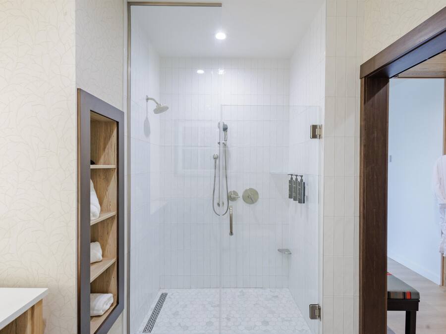 A modern bathroom with a shower, a wooden shelf with towels, and a white tiled floor.