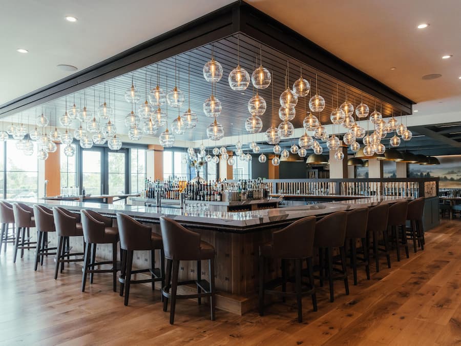 Een bar met een lange toonbank, meerdere krukken en hangende bolvormige lampen.