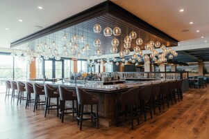 Zona de bar con una barra alargada, varios taburetes e lámparas esféricas colgantes.