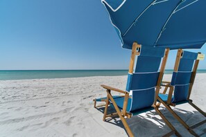 A beach chair and umbrella setup on a sandy beach with a clear blue sky. - Coastal Snowbird Haven: Beach, Golf, Tennis & Pool (Destin)
