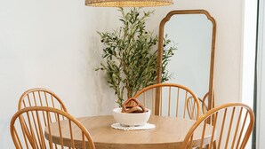 Une table de salle à manger ronde en bois, accompagnée de quatre chaises, une suspension en fibres tressées et une plante en pot en arrière-plan.