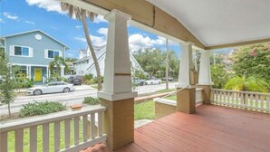 Une véranda couverte avec des balustrades blanches et une terrasse en bois, donnant sur une rue bordée de maisons et de voitures.