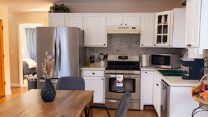 Cocina moderna con armarios blancos, un refrigerador de acero inoxidable, un microondas y una estufa. Hay una mesa de comedor con sillas y un jarrón de flores secas.