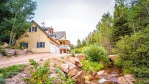 Una casa amarilla de dos pisos con un balcón, rodeada por un paisaje natural con un arroyo y árboles frondosos.