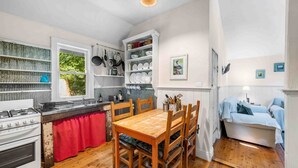 Cocina compacta con estufa a gas, mesa de madera y sillas, zona de comedor con mantel individual rojo, y un dormitorio con dos camas.